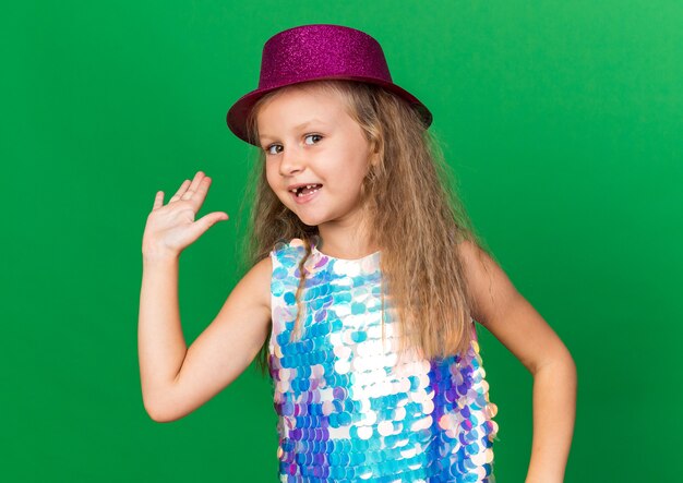 コピースペースと緑の壁に分離された上げられた手で立っている紫色のパーティハットと笑顔の小さなブロンドの女の子