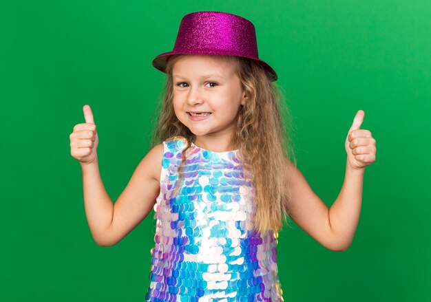 Free photo smiling little blonde girl with purple party hat thumbing up isolated on green wall with copy space