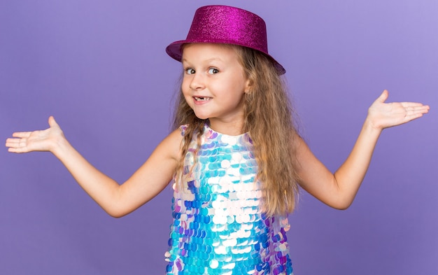 Smiling little blonde girl with violet party hat holding hands open isolated on purple wall with copy space