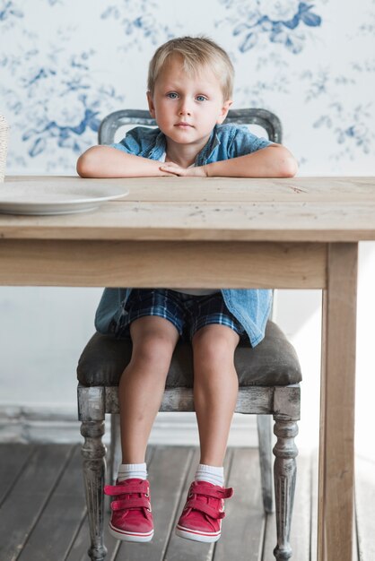 Улыбаясь маленький мальчик, сидя на стуле перед деревянным обеденным столом
