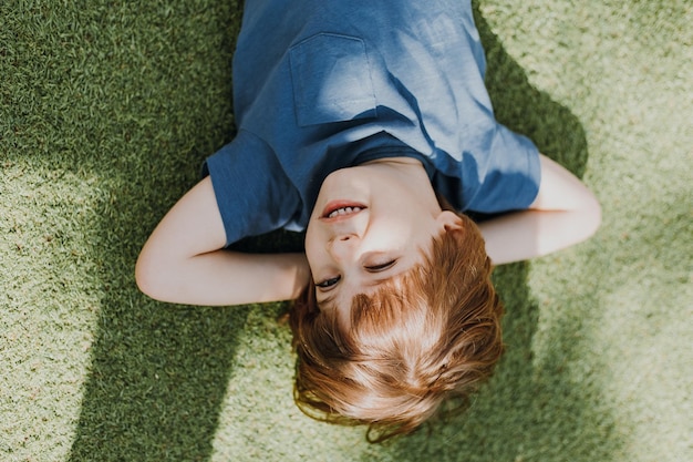Smiling little brunette boy in a blue t-shirt lies on the green grass with his hands under his head. child is resting outdoors. kid takes air baths. lifestyle. space for text. top view