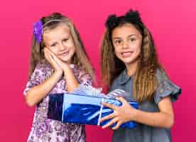 Free photo smiling little caucasian girl holding gift box and standing with pleased little blonde girl isolated on pink wall with copy space