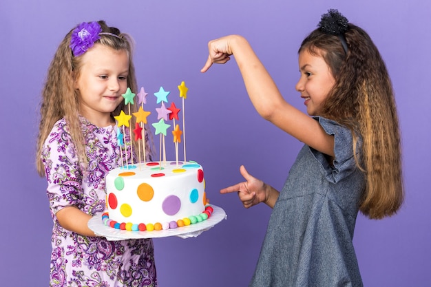 Free photo smiling little caucasian girl pointing at pleased little blonde girl holding birthday cake isolated on purple wall with copy space