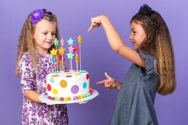 コピースペースで紫色の壁に分離されたバースデーケーキを保持している幸せな小さなブロンドの女の子を指して笑顔の小さな白人の女の子