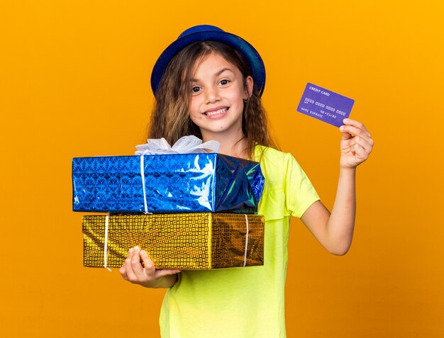 コピースペースとオレンジ色の壁に分離されたクレジットカードとギフトボックスを保持している青いパーティーハットを持つ白人の少女