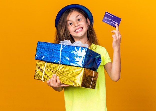 smiling little caucasian girl with blue party hat holding gift boxes and credit card isolated on orange wall with copy space