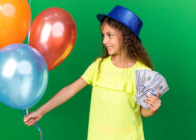 Smiling little caucasian girl with blue party hat holding money and looking at helium balloons isolated on green wall with copy space