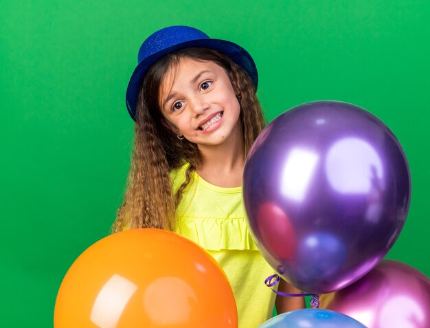 улыбающаяся маленькая кавказская девочка в синей партийной шляпе держит гелиевые шары, изолированные на зеленой стене с копией пространства