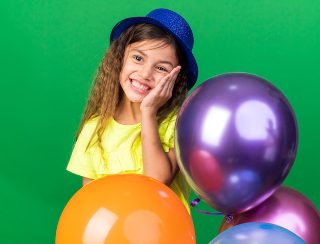 블루 파티 모자 얼굴에 손을 넣고 복사 공간이 녹색 벽에 고립 된 헬륨 풍선을 들고 웃는 어린 백인 소녀