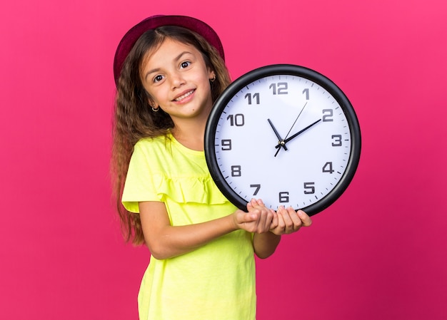 smiling little caucasian girl with purple party hat holding clock isolated on pink wall with copy space