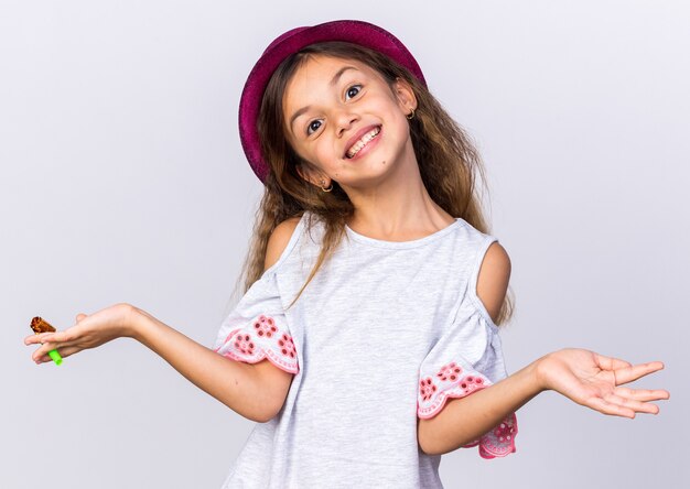 smiling little caucasian girl with purple party hat holding party whistle and keeping hand open isolated on white wall with copy space