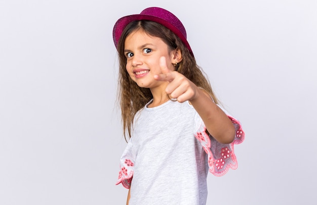 smiling little caucasian girl with purple party hat pointing  isolated on white wall with copy space