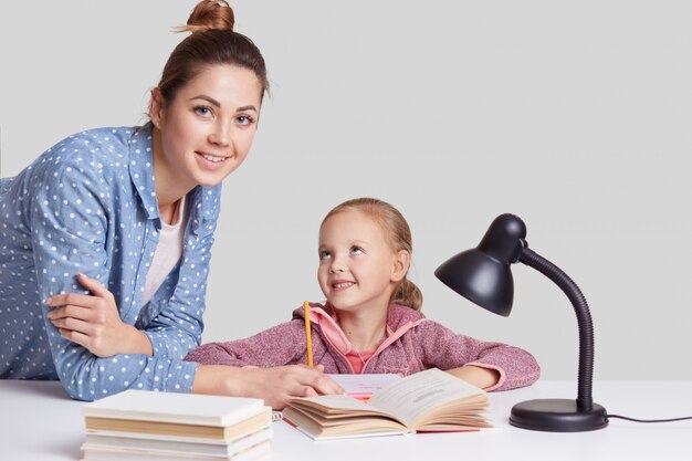 Smiling little charming girl sits at table, does homework task together with her mother, try to write composition, look joyfully, uses reading lamp for good vision, isolated on white wall