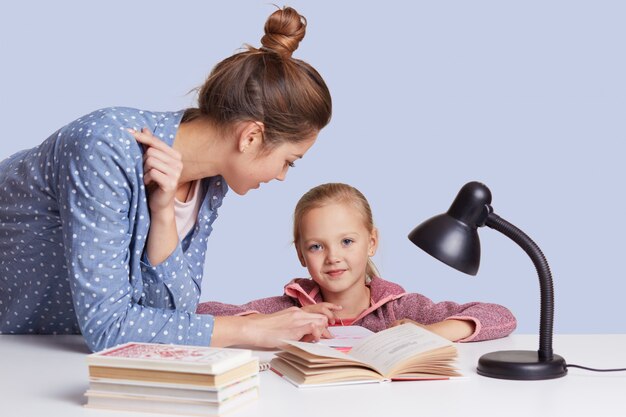 Smiling little charming girl sits at table and her mother help her to do homework task, try to learnn poem together, uses reading lamp for good vision, isolated on white.