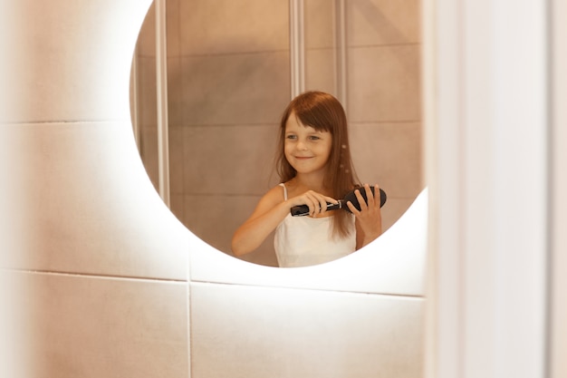 Улыбающаяся маленькая девочка расчесывает волосы в ванной перед зеркалом, любит смотреть на свое отражение, носить домашнюю одежду, делать косметические процедуры.