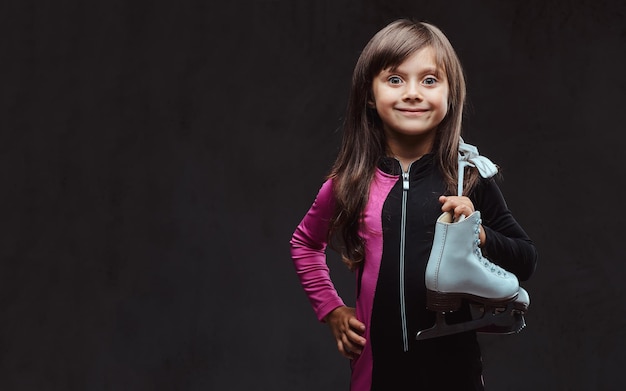 Smiling little girl dressed in sportswear holds ice skates on a shoulder. Isolated on a dark textured background.