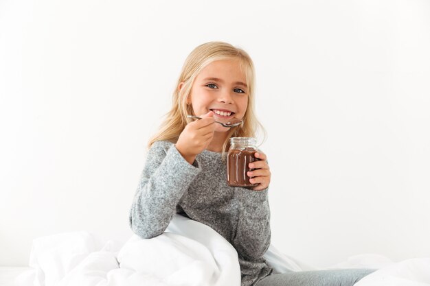 Smiling little girl holding bank of sweet chocolate hazelnut spread,  while sitting in bed