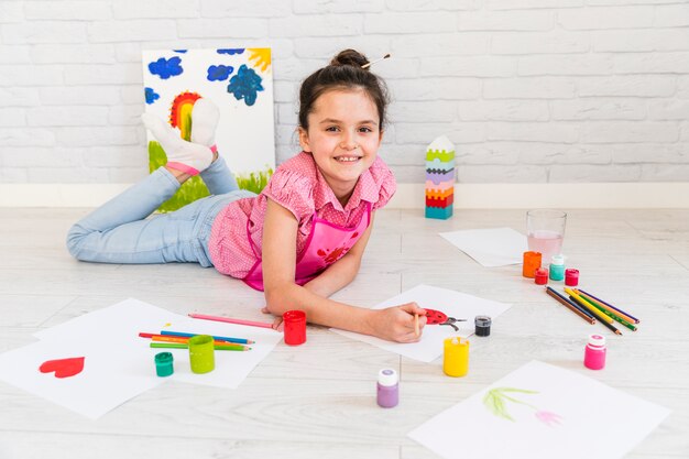 Smiling little girl lying on floor painting with paint brush on white paper