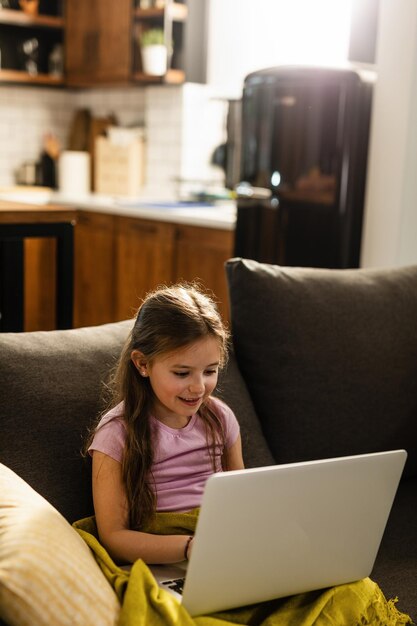 소파에 앉아 거실에서 노트북을 사용하는 웃는 어린 소녀