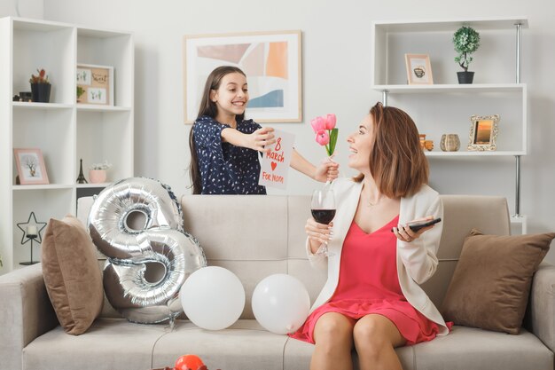 Smiling little girl standing behind sofa gives frowers with postcard to smiling mom with phone and glass of wine in living room