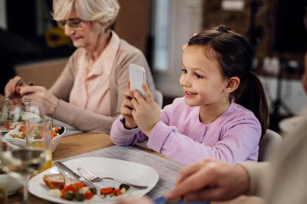 Smiling little girl using mobile phone while sitting at dining table during lunch time