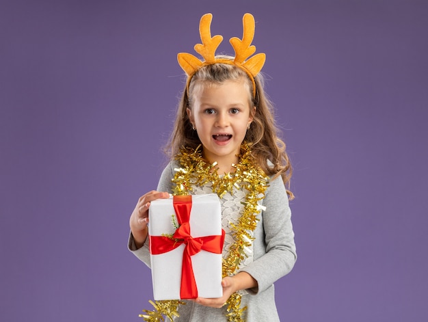 smiling little girl wearing christmas hair hoop with garland on neck holding gift box isolated on blue wall