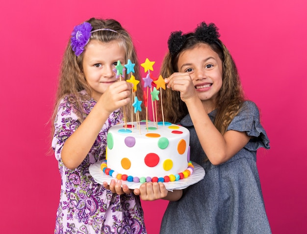 smiling little pretty girls holding birthday cake together isolated on pink wall with copy space