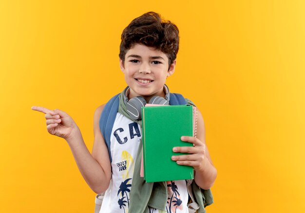 コピースペースで黄色に隔離された側に本とポイントを保持しているバックバッグとヘッドフォンを身に着けている小さな男子生徒の笑顔