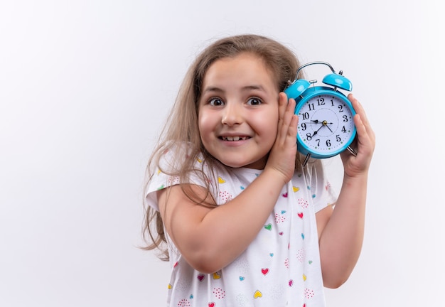 Smiling little school girl wearing white t-shirt holding alarm clock around ear on isolated white background