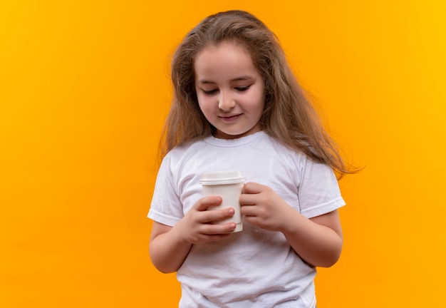 Улыбающаяся маленькая школьница в белой футболке держит чашку кофе на изолированном оранжевом фоне