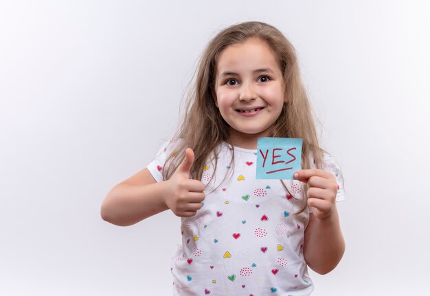 紙を保持している白いTシャツを着て笑顔の小さな女子高生は、孤立した白い背景に親指をマーク