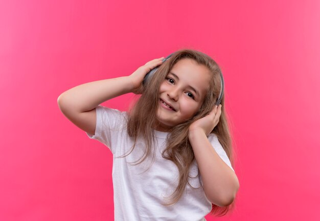 Улыбающаяся маленькая школьница в белой футболке слушает музыку в наушниках на изолированном розовом фоне
