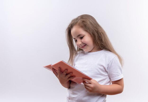 Smiling little school girl wearing white t-shirt looking at notebook on isolated white background