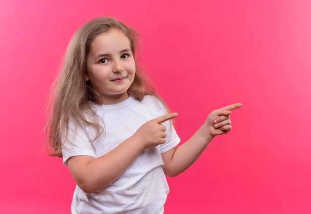 Smiling little school girl wearing white t-shirt points to side on isolated pink background