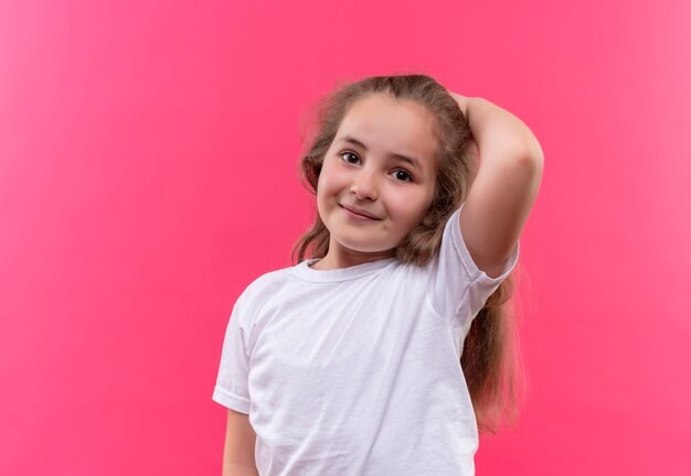Улыбающаяся маленькая школьница в белой футболке положила руку на голову на изолированном розовом фоне