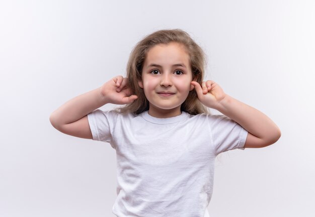 Улыбающаяся маленькая школьница в белой футболке положила руки на уши на изолированном белом фоне