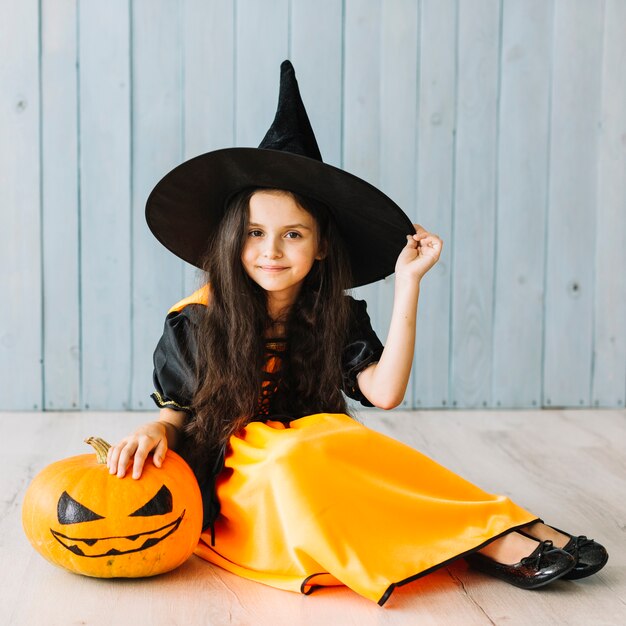 Smiling little witch sitting with pumpkin on floor