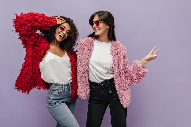 Smiling long haired lady with red sunglasses in white top dark pants and pink sweater smiling together with mulatto curly girl in bright clothes