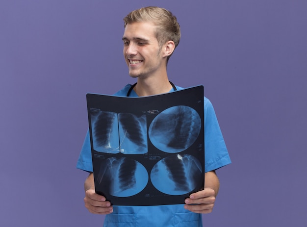 Улыбаясь, глядя в сторону, молодой мужчина-врач в униформе врача со стетоскопом, держащим рентгеновский снимок, изолированный на синей стене