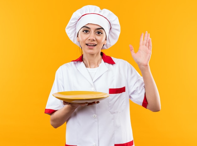 Smiling looking at camera young beautiful girl in chef uniform holding plate raising hand 