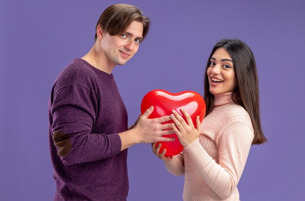 Улыбаясь, глядя в камеру, молодая пара в день святого валентина держит воздушный шар сердца, изолированные на синем фоне