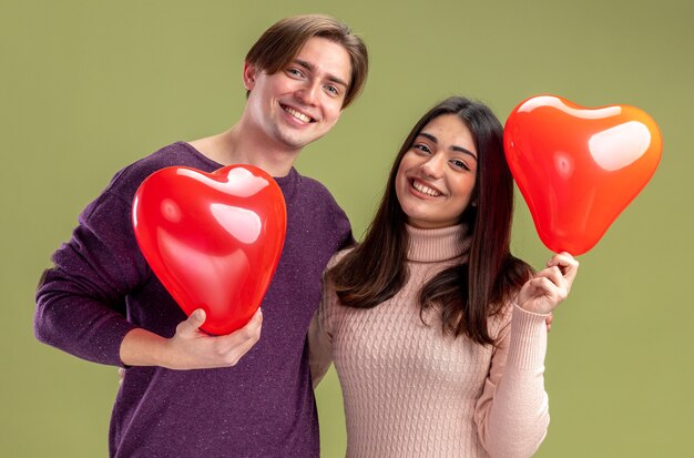 Улыбаясь глядя камеру молодая пара в день святого валентина держит воздушные шары сердца, изолированные на оливково-зеленом фоне