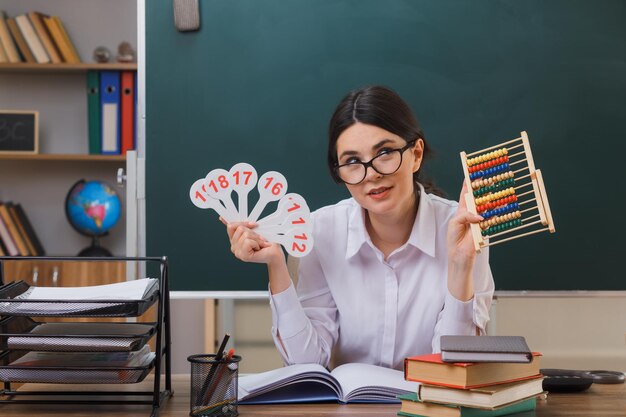 улыбаясь, глядя в камеру, молодая учительница в очках держит счеты с числовым вентилятором, сидя за партой со школьными инструментами в классе