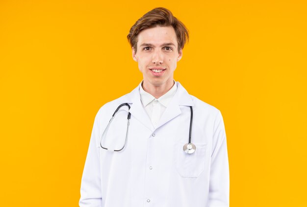 Smiling looking camera young male doctor wearing medical robe with stethoscope isolated on orange wall