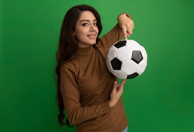 Улыбаясь, глядя на переднюю молодую красивую девушку, держащую мяч в пальце, изолированную на зеленой стене
