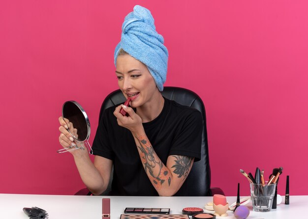 Smiling looking at mirror young beautiful girl sits at table with makeup tools wrapped hair in towel applying lipstick isolated on pink background