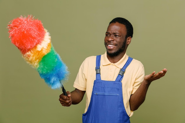 Smiling looking at side holding pipidaster young africanamerican cleaner male in uniform isolated on green background