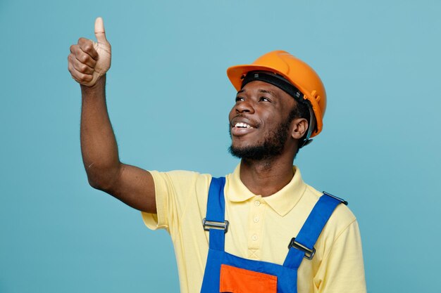 Smiling looking at side showing thumbs up young african american builder in uniform isolated on blue background