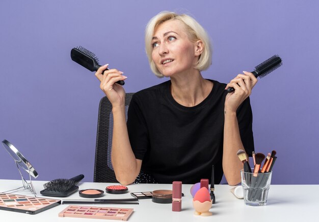 Smiling looking side young beautiful girl sits at table with makeup tools holding combs isolated on blue background