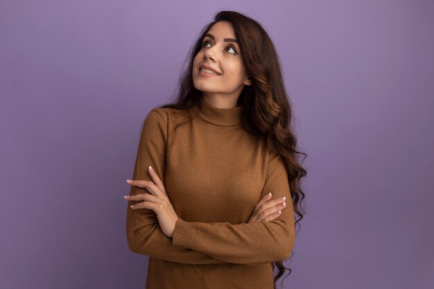 Smiling looking at side young beautiful girl wearing brown turtleneck sweater crossing hands isolated on purple wall